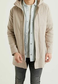 Man som bär en beige zip-up jacka över en ljusblå skjorta med knappar och en vit T-shirt, ihop med mörkgrå jeans.