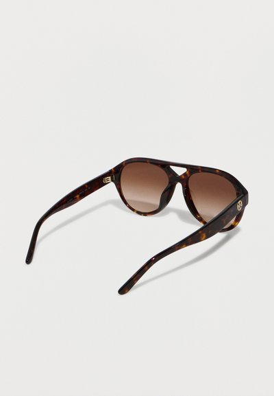 Γυαλιά ηλίου τύπου αεροπόρου με καφέ φακούς με διαβάθμιση, γέφυρα σε σχήμα κλειδαρότρυπας και χρυσό λογότυπο στο πλαϊνό μέρος, τοποθετημένα σε λευκή επιφάνεια.