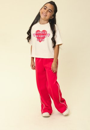 Lachend meisje met lang donker haar, draagt een wit T-shirt met "Lonely Hearts Club" bedrukt, rode broek met witte strepen en witte sneakers.