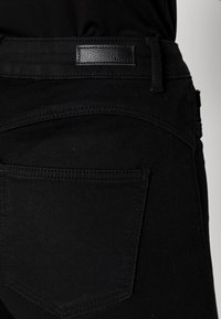 Jean en denim noir avec une texture lisse, un design à cinq poches et une petite étiquette en cuir sur la ceinture. Des détails de couture subtils soulignent la forme.