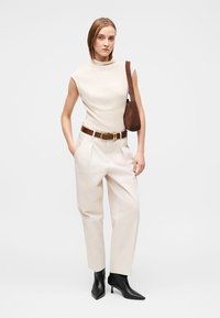 Top creme sem mangas com gola alta, calças brancas de cintura alta, cinto castanho, mala castanha e botins pretos com bico fino. Design simples e estruturado.