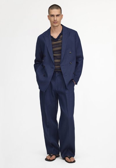 Modèle masculin debout, portant un costume marine ample croisé, un haut en maille rayé et des tongs noires sur un fond blanc.