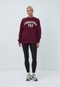 Burgunderfarget sweatshirt med teksten "LEGERSTUDIO", oversized passform, ribbede mansjetter, kombinert med svarte leggings og treningssko. Enfarget bakgrunn.