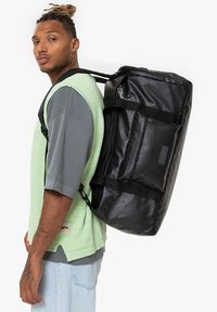 Musta reppu kiiltävällä pinnalla, jossa on useita taskuja, säädettävät hihnat ja sylinterimäinen muoto, kuljetettuna mintunvihreän paidan päällä.