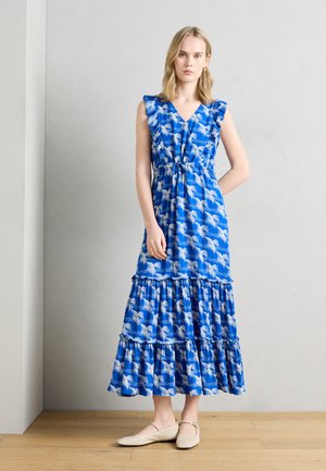 Γυναίκα που φοράει μπλε μακρύ φόρεμα χωρίς μανίκια με λευκό σχέδιο από φύλλα φοίνικα και μπεζ φλατ παπούτσια, στέκεται μπροστά σε ανοιχτό γκρι τοίχο και ξύλινο πάτωμα.