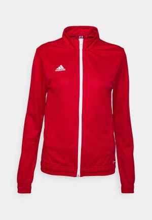 Crvena atletska jakna napravljena od glatkog, laganog materijala. Ima visoki ovratnik, punu patent zatvarač i bijeli Adidas logo na lijevoj strani prsnog koša.