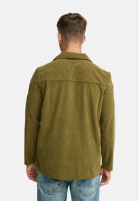 Oliven grøn kordfløjls skjortejakke med krave, lange ærmer og løs pasform. Har vertikal ribbet tekstur og bæres med lyseblå jeans.