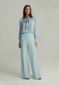 Helles blaues Blusenshirt mit blumigen Mustern und Schleifenverschluss, kombiniert mit hoch taillierten, weit geschnittenen hellblauen Hosen. Glatte Stoffbeschaffenheit.