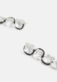 Jasni kristalni uhani z vrsto kvadratnih kamnov, nastavitvenih v črnem kovinskem okviru, s poliranim srebrnim kovinskim poudarkom in zapiranjem na zamašek.