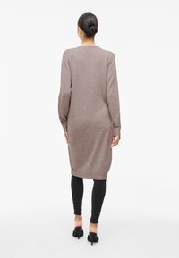 Kvinna med mörkt hår i knut bär en lång mauvefärgad klänning, svarta leggings och svarta klackskor, stående vänd bort från kameran mot vit bakgrund.