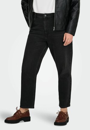 Asmuo dėvi juodus džinsus, rudos odinės suvarstomos batelius, baltą marškinių ir juodą odinę striukę priešais paprastą foną.