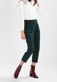 Blusa bianca a maniche lunghe con collo ricamato, abbinata a pantaloni verde scuro a vita alta e stivaletti glossy bordeaux.