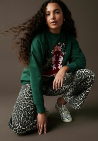 Zelená mikina s vianočným motívom, skombinovaná s nohavicami s leopardím vzorom. Modelka má na sebe strieborné tenisky a dlhé kučeravé vlasy.