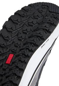 Suela de zapato de goma negra con surcos profundos para tracción, mediopie en blanco y parte superior texturizada en gris. Presenta un acento rojo y la etiqueta "ALL-TRACK".