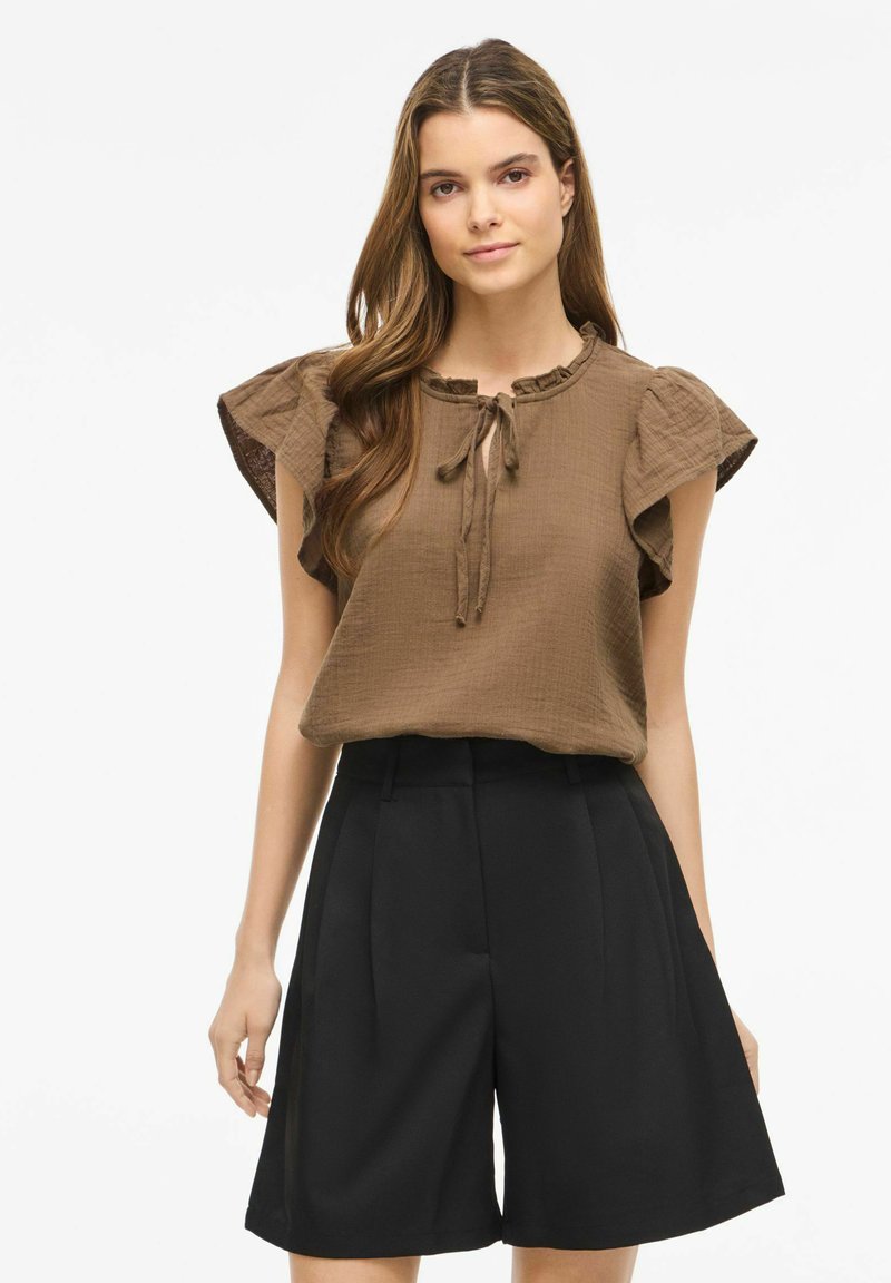 Jeune femme aux longs cheveux bruns portant un chemisier texturé marron à manches courtes avec un nœud au col et un short noir taille haute à jambes larges.