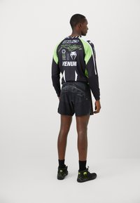 Langärmliges schwarzes Sportoberteil mit grünen Akzenten und Logos, kombiniert mit schwarzen Shorts mit Grafikdruck. Getragen mit schwarzen Turnschuhen.