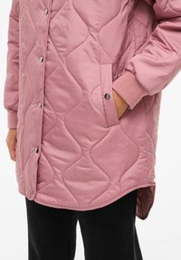 Odebeljena roza zunanji plašč z zatičnimi gumbi, stranskimi žepi in rebrastimi manšetami. Ima rahlo ukrivljen rob za sproščen oprijem.