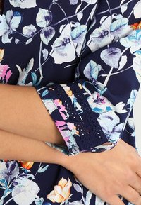 Blusa navy con motivo floreale che presenta fiori blu, rosa e bianchi. Maniche decorate con pizzo e chiusura a bottone. Tessuto morbido.