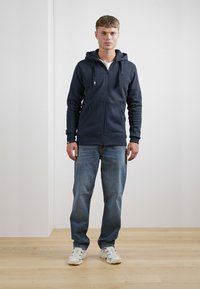 Mørkeblå zip-up hoodie med snøre, frontlommer, parret med lys udfadede blå jeans og hvide sneakers. Enkel design, afslappet pasform.
