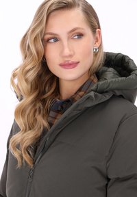 Szőke, hullámos hajú nő ezüst fülbevalót visel, zöld, kapucnis párnázott dzsekit és alatta egy barna kockás inget hord, oldalra néz.