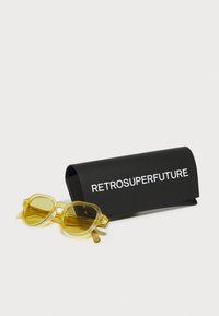 Žute prozirne sunčane naočale s oktagonalnim okvirom, odložene pored crne futrole s bijelim tekstom "RETROSUPERFUTURE". Mat tekstura.