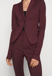 Blazer aderente bordeaux con chiusura a due bottoni, rever a punta, tasche laterali e una struttura ben definita, abbinato a pantaloni sartoriali coordinati.