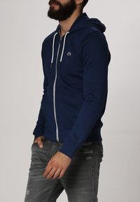 Marinblå zip-up hoodie tillverkad av bomullsblandat tyg, med en framficka, dragsko i huvan och en liten grön logotyp på bröstet.