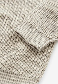 Stickad tröjsleeve i ljusbeige med en texturerad, ribbad mönstring. Har en mjuk, vävd yta och synliga sömnadsdetaljer.