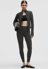 Conjunto deportivo que incluye una chaqueta negra con puntos y leggings a juego, ambos hechos de tejido elástico, con una esterilla de yoga enrollada debajo de un brazo.
