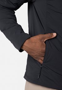 Zwarte, lichtgewicht jas met een gestructureerde afwerking, voorzien van een zijzak en een verstelbare zoom met een koord. Arm zichtbaar.