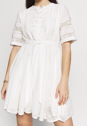 Donna che indossa un vestito bianco con pizzo sui bordi, bottoni sul davanti e cintura annodata in vita.