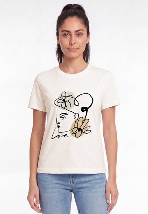 Femme aux cheveux foncés portant un t-shirt blanc avec un dessin au trait noir d'un visage, des fleurs et le mot "Love", debout sur fond blanc.