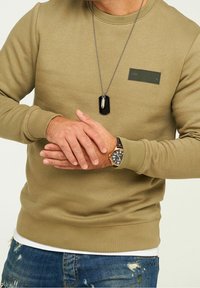 Uomo in felpa verde oliva, orologio da polso nero e oro, collana con dog tag e jeans blu strappati in posa casual.