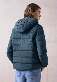 Giacca trapuntata verde acqua con cuciture orizzontali, cappuccio e chiusura con zip. Il tessuto appare liscio e leggero. Presenta una vestibilità rilassata.
