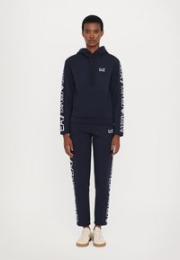 Conjunto de sudadera y pantalones de chándal en color navy, fabricado en mezcla de algodón. La sudadera cuenta con un bolsillo delantero y un logo, mientras que los pantalones presentan detalles del logo en los laterales. Zapatos ligeros.
