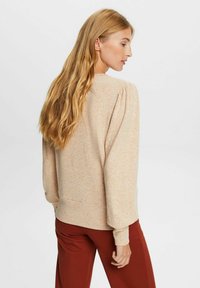 Suéter de color beige con punto texturizado, mangas abullonadas y puños de canalé. Combinado con pantalones de color óxido, visto desde la parte de atrás.