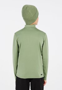 Camicia verde a maniche lunghe con colletto alto, dalla texture liscia, con un'etichetta nera in basso, abbinata a un berretto verde a coste coordinato.