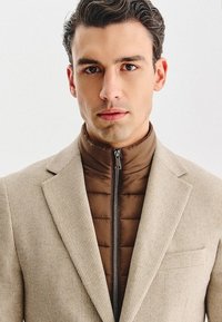 Mężczyzna z ciemnymi włosami, ubrany w beżową marynarkę na brązowej pikowanej kurtce zapinanej pod szyję, zwrócony przodem z neutralnym wyrazem twarzy.