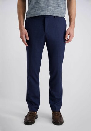 Mann trägt marineblaue Anzughose, graues T-Shirt und dunkelbraune Leder-Loafer, steht vor einem schlichten hellen Hintergrund.