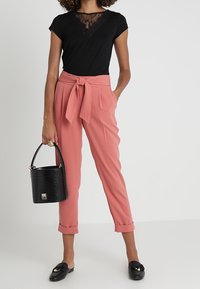 Camiseta negra con detalle de encaje, pantalones rosa con cinturón anudado, bolso negro y mocasines negros. La tela es suave con un ajuste estructurado.