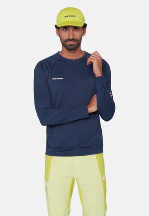 Homme portant une casquette jaune Mammut, une chemise à manches longues bleu marine avec le logo Mammut, et un pantalon jaune avec des poches latérales, debout devant un fond blanc.