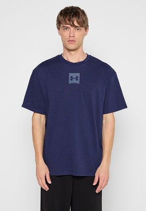 Młody mężczyzna stojący przodem, ubrany w gładką, ciemnoniebieską koszulkę Under Armour i czarne spodnie na białym tle.