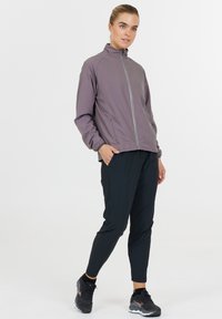 Let let jakke i mauve med lynlås, ståkrave og elastiske manchetter, parret med sorte tapered bukser og sorte sportsko.
