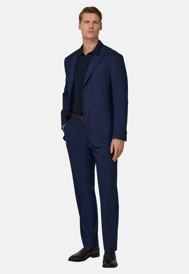Homme debout portant un costume bleu marine, une chemise noire, une ceinture marron et des chaussures noires, sur un fond clair uni.