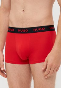 Boxer in cotone rosso con elastico nero e scritta "HUGO" in lettere rosse. Il design è aderente e presenta una superficie liscia.