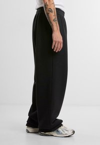 Melni, vaļīgi sporta bikses ar elastīgu jostu. Mīksts audums, šauras piedurknes un kabatas. Valkātas ar pelēkiem un zilajiem sporta apaviem.