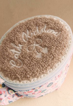 Okrągła saszetka z beżową, miękką górą, wyszywaną napisem „Cat & Nutmeg”, oraz korpusem z materiału w kwiatowy wzór, zapinana na zamek i wyposażona w materiałową pętlę.