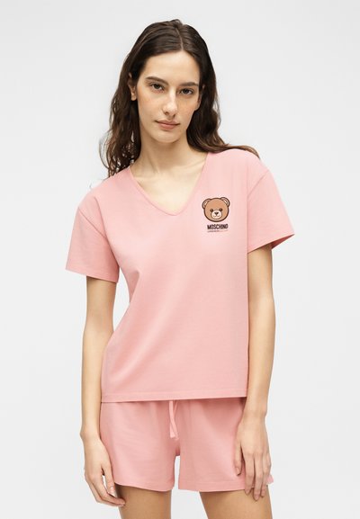 Γυναίκα φοράει ροζ κοντομάνικο τοπ με V λαιμόκοψη και αντίστοιχα σορτς με λογότυπο αρκουδάκι και κείμενο "Moschino Underwear" στο στήθος.