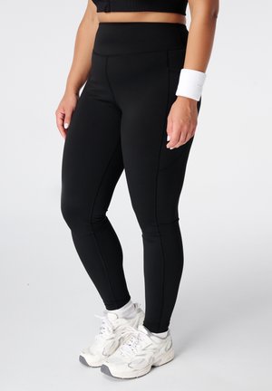 Personne portant un legging noir taille haute, des baskets blanches, des chaussettes blanches et un bracelet blanc au poignet droit sur un fond uni.