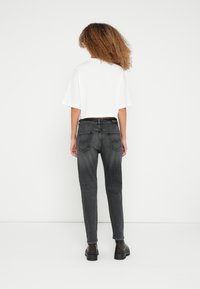 Göndör hajú személy háttal áll, fehér, rövid felsőt, sötétszürke, magasderekú farmert és fekete, masszív bakancsot visel.